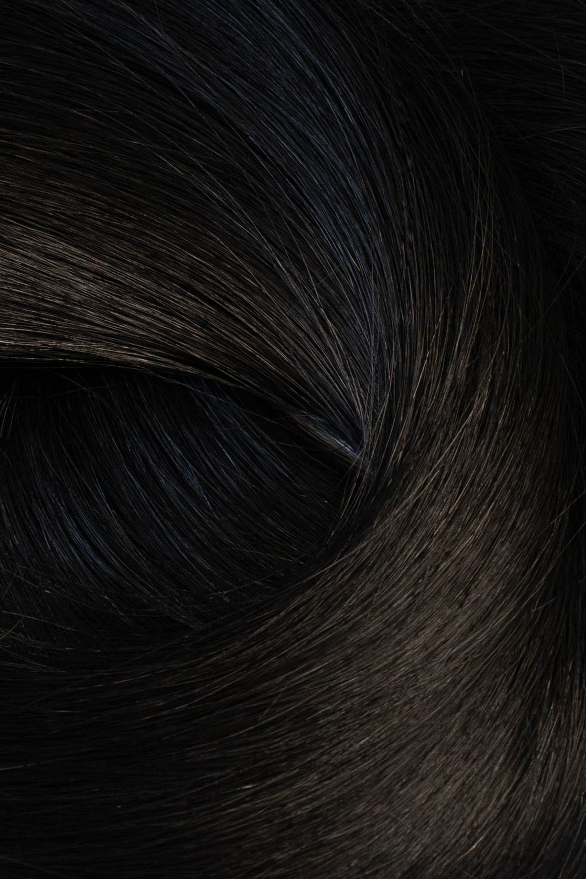 A close-up texture shot of dark, glossy hair with subtle warm brown undertones and a soft, swirled arrangement. (Jet Black / #1)