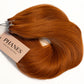 A bundle of PHANES hair extension wefts in a warm coppery auburn shade, with a beige paper tag attached, photographed on a clean white background. (Cowgirl Copper / #180)