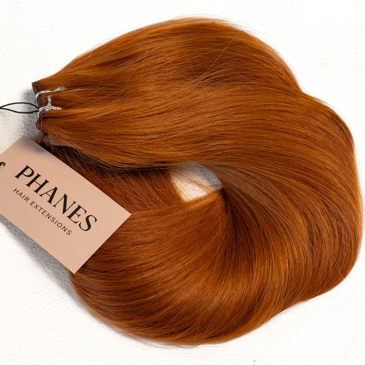 A bundle of PHANES hair extension wefts in a warm coppery auburn shade, with a beige paper tag attached, photographed on a clean white background. (Cowgirl Copper / #180)