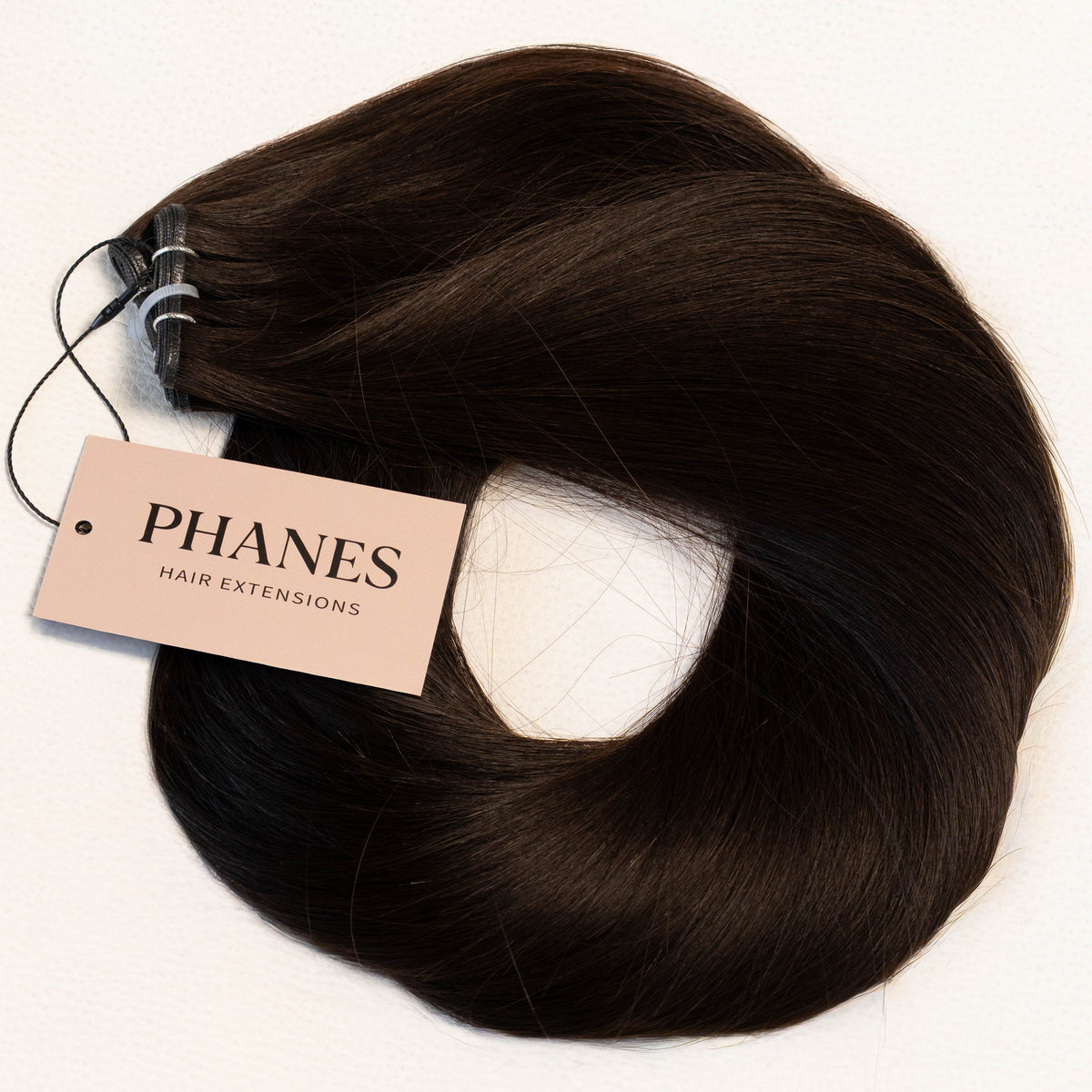 A bundle of PHANES hair extensions in a deep dark brown color, coiled on a white background with a brand tag attached. (Soft Black / #1b)