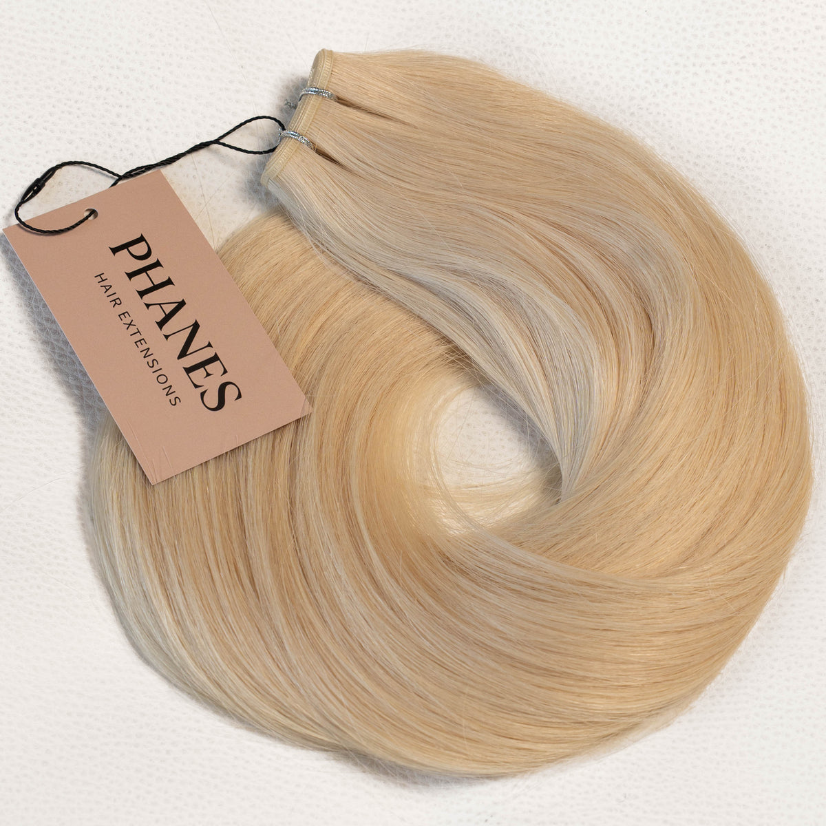 A bundle of blonde hair extensions with a light brown paper tag reading PHANES, arranged in a loose coil on a white textured background. (Ice Blondest / #24A)