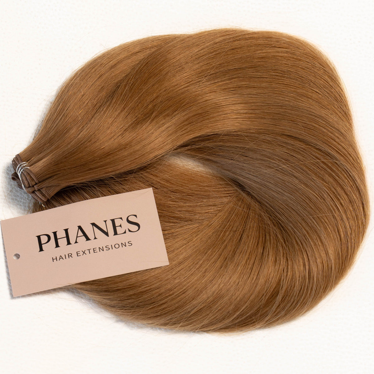 A bundle of PHANES hair extensions in a warm auburn-brown shade, arranged in a loose curl with a beige brand tag attached, on a plain white background. (Caramel / #8)