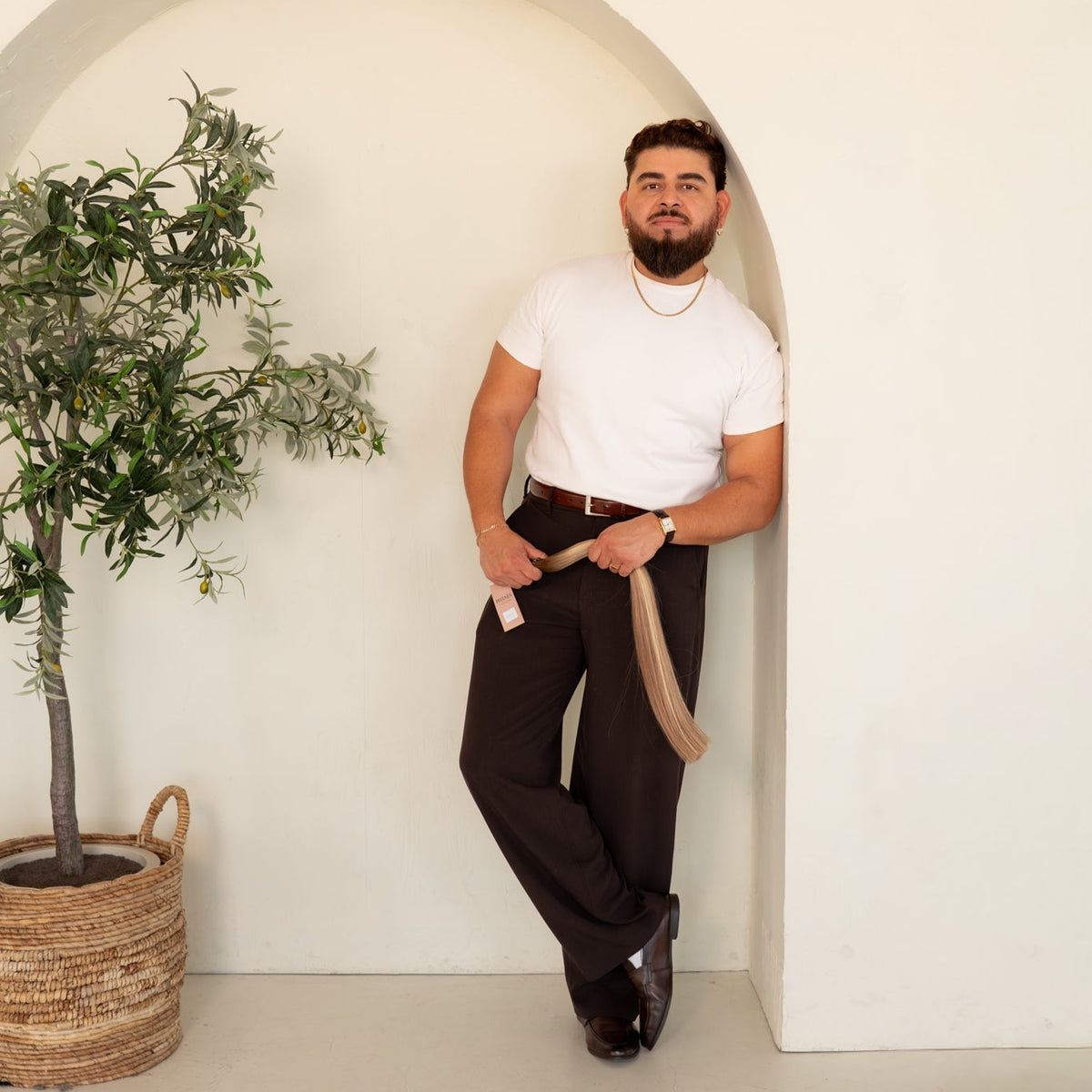 A man with a beard wearing a white t-shirt, dark brown trousers, and a beige belt, posing in front of a white wall with a decorative arch and a potted plant.