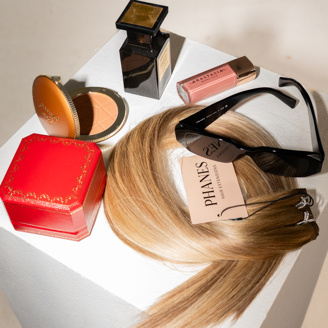 A styled flat-lay of multiple luxury beauty products and a hair extension on a white pedestal. The set includes a Phlanes blonde hair extension, a Tom Ford compact powder, a Tom Ford perfume bottle, a Tom Ford lipstick, a red Cartier box, and black sunglasses.