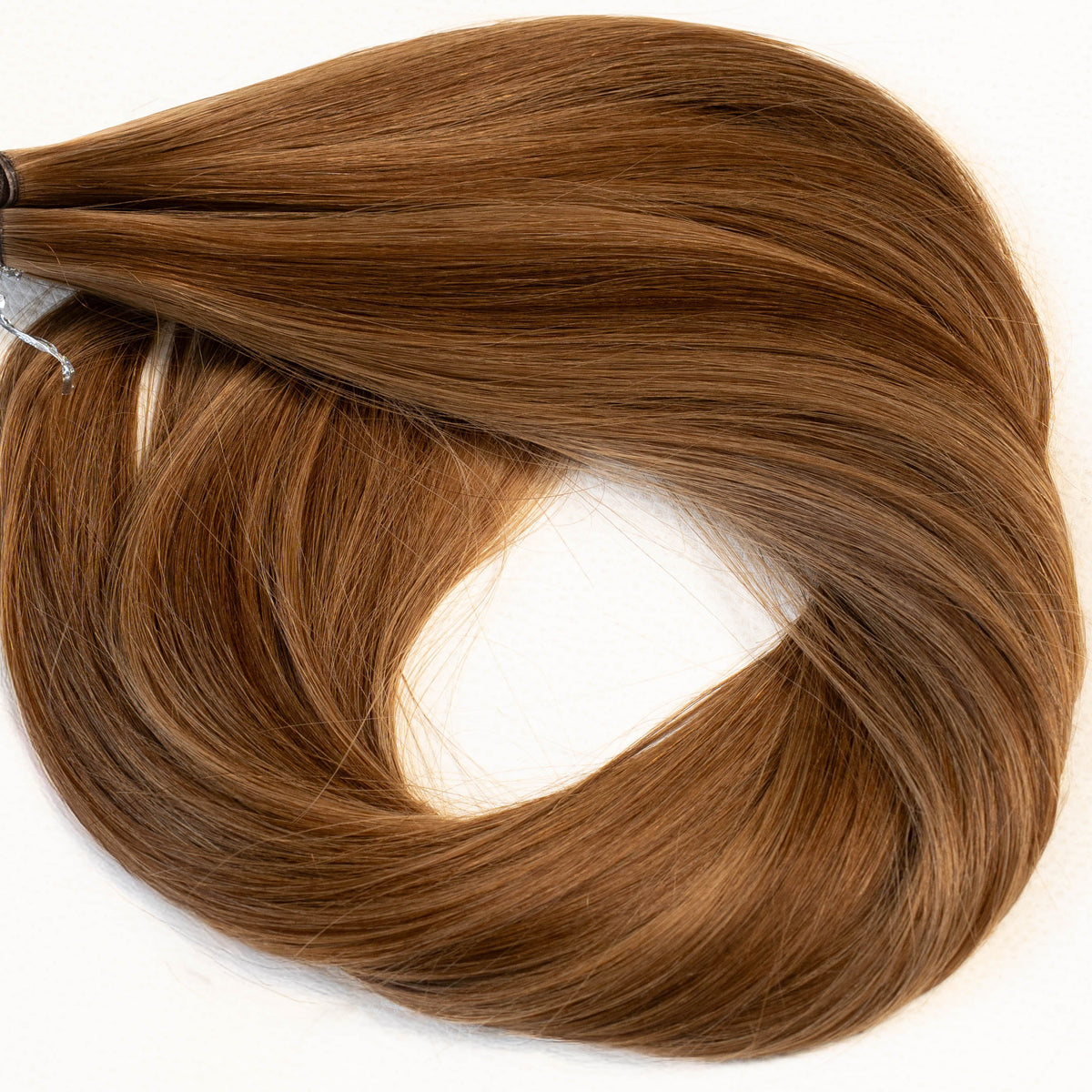 A bundle of medium brown hair extension pieces with a small gray attachment tab, arranged in a loop on a plain white background. (Caramel Blonde / #P4/8)