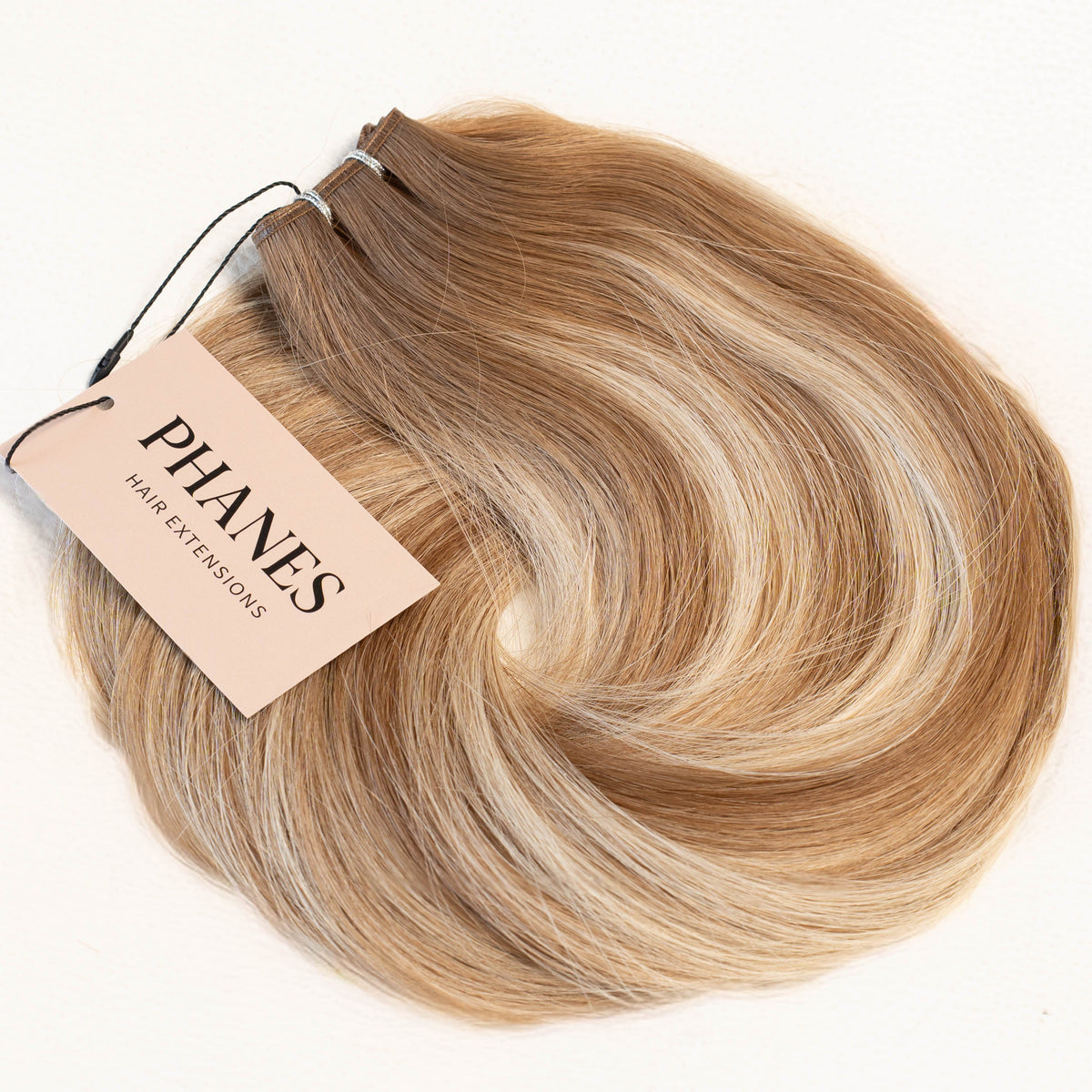 A bundle of Phanes hair extensions in a light brown and blonde ombré color, with a beige paper tag attached, photographed on a clean white background. (Malibu Balayage / T#8A-P8A/1001)
