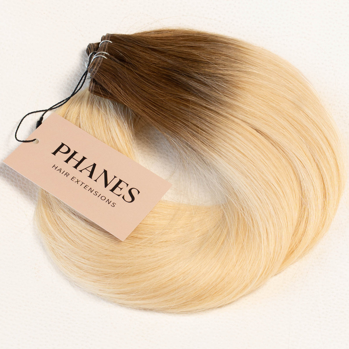 A bundle of PHANES hair extensions with a brown to blonde ombré color, featuring a light pink paper tag attached, set against a plain white background. (Rooted Blonde / #T4/60)
