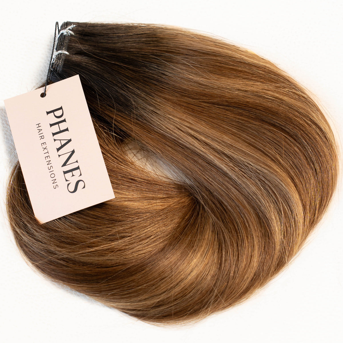 A hair extension bundle with a tan tag reading PHANES, showing a warm brown to blonde ombré color, photographed on a solid white background. (Carmel Mocha / TBF)