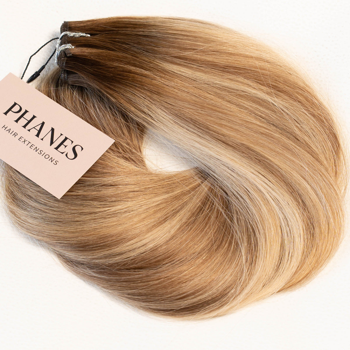 A bundle of synthetic hair extensions with a light pink tag reading 'PHANES HAIYI EXTENSION', featuring a blonde-to-brown ombré color gradient, set against a plain white background. (Brooklyn Balayage / #TFF)