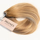 A bundle of Phanes hair extensions in a light golden blonde shade with subtle warm brown lowlights, featuring a pink paper tag with the brand name 'PHANES' on a clean white background. (Miami Balayage / TLQ)