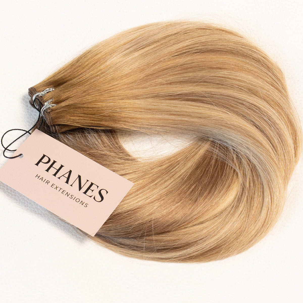 A bundle of Phanes hair extensions in a light golden blonde shade with subtle warm brown lowlights, featuring a pink paper tag with the brand name 'PHANES' on a clean white background. (Miami Balayage / TLQ)