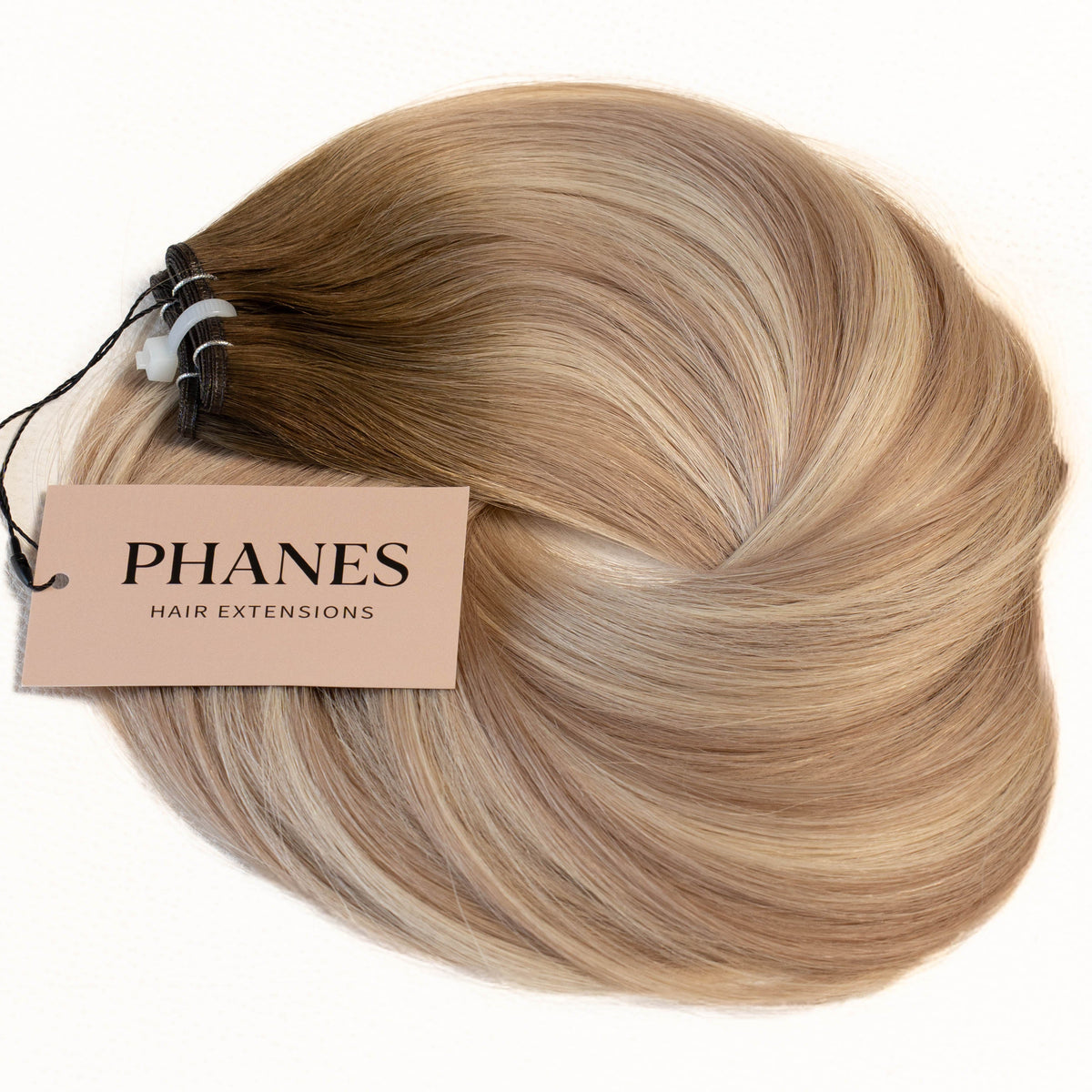 A bundle of Phanes hair extensions in a light blonde shade with subtle warm brown undertones, featuring a paper brand tag on a clean white background. (Icy Balayage / #TSK)