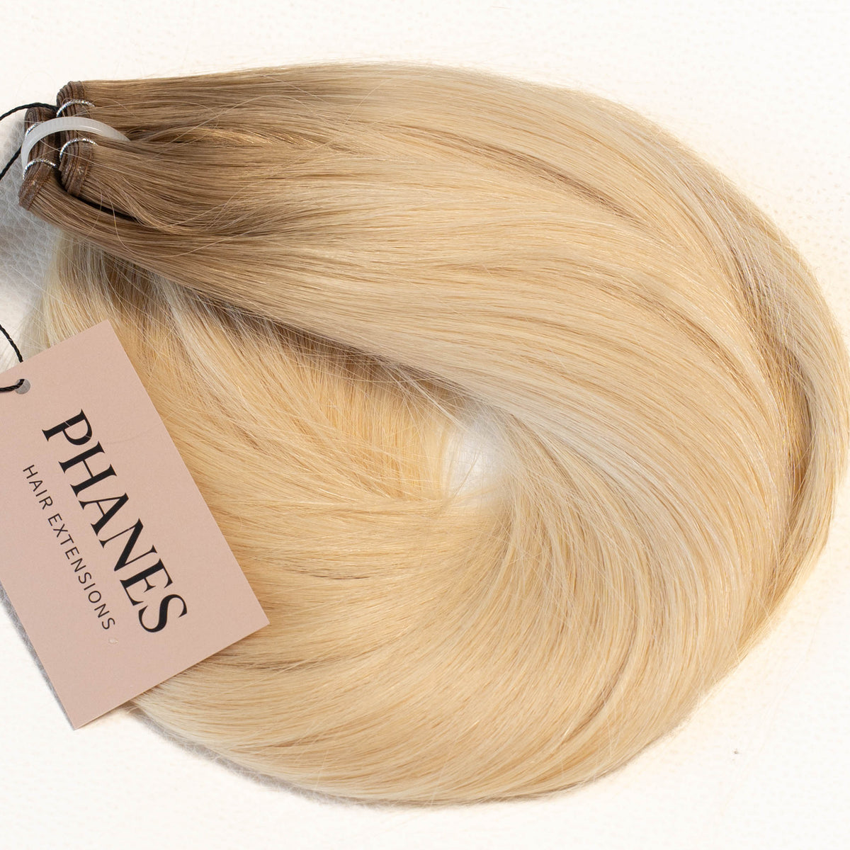 A bundle of Phanes hair extensions in a blonde ombré color, featuring a light beige to golden blonde gradient, with a pink brand tag attached on a plain white background. (Cool Ombré / #TTA)