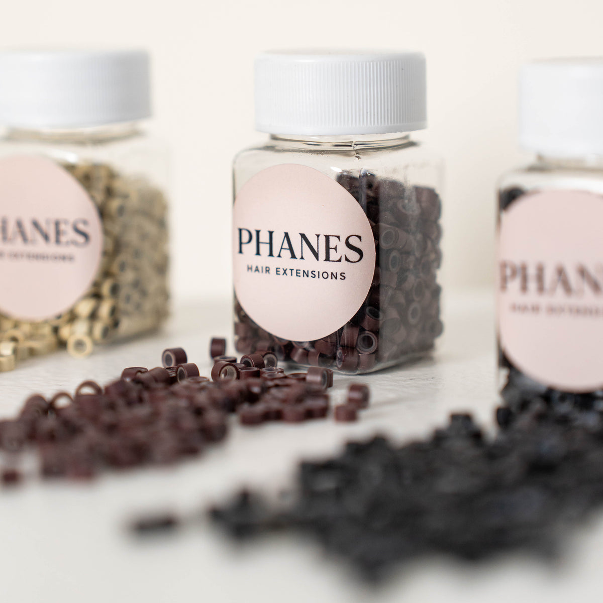 Three clear plastic jars of PHANES hair extensions with white lids and pink labels, arranged on a light surface with loose dark brown and black extension pieces scattered in front.