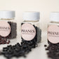 Three clear plastic jars of PHANES Hair Extensions with white caps and pink labels, with loose black hair extension beads spilled in front on a light surface.