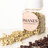 A clear bottle labeled PHANES HAIR EXTENSIONS with a white cap, filled with small beige ring-shaped hair extension pieces and spilled onto a white surface, with some dark brown pieces in the foreground. (Blonde)