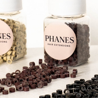 Three clear plastic bottles of PHANES hair extensions with white ribbed caps, showing beige, dark brown, and black extension pieces inside. Loose pieces of the same extensions are scattered on a white surface in front of the bottles. (Brown)