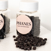 Clear plastic jars of PHANES Hair Extensions filled with small black ring-shaped hair extension beads, with some beads spilled in front on a white surface. (Dark Brown)