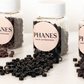 Several clear plastic bottles of PHANES hair extensions with white ribbed caps and round pale pink labels, with loose black hair extension pieces spilled in front on a white surface. (Black)