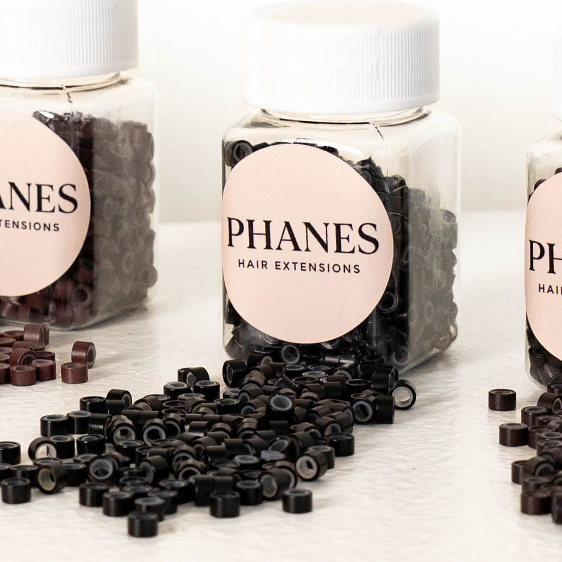 Several clear plastic bottles of PHANES hair extensions with white ribbed caps and round pale pink labels, with loose black hair extension pieces spilled in front on a white surface. (Black)