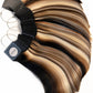 A fan-shaped bundle of hair extensions or wigs with layered blonde, brown, and black streaks, attached to black clips with metal rings, photographed on a plain white background.