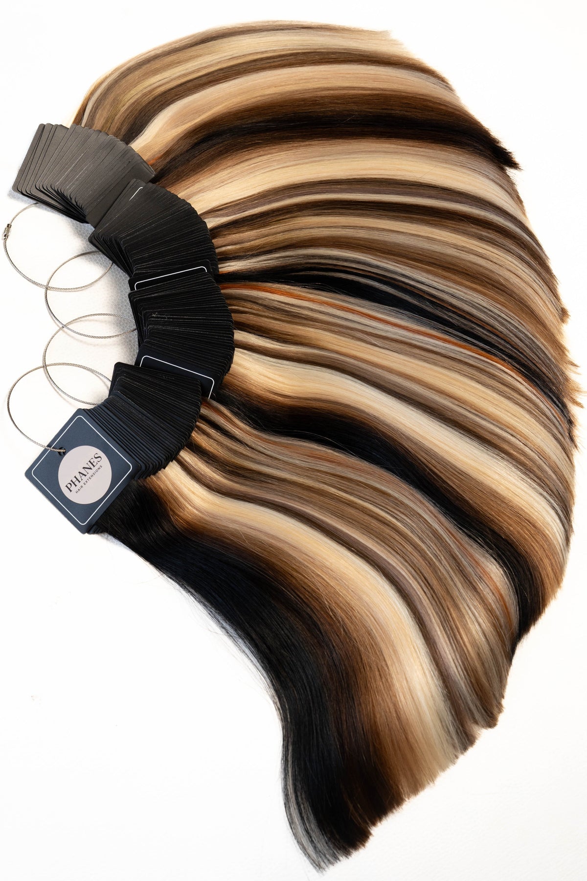 A fan-shaped bundle of hair extensions or wigs with layered blonde, brown, and black streaks, attached to black clips with metal rings, photographed on a plain white background.